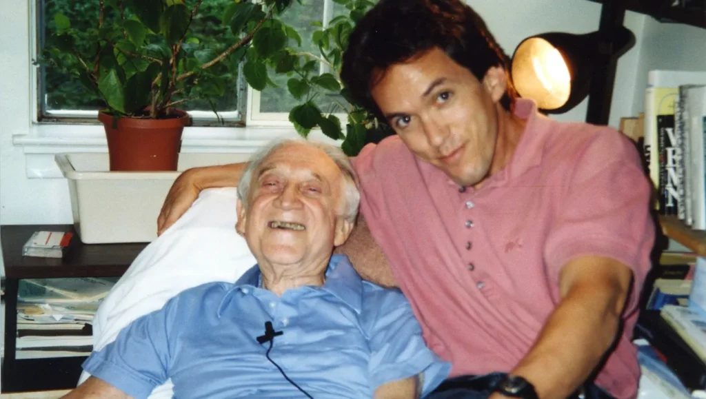 Mitch Albom sitting beside Morrie Schwartz, sharing a heartfelt conversation about life-changing books during one of their Tuesday visits.