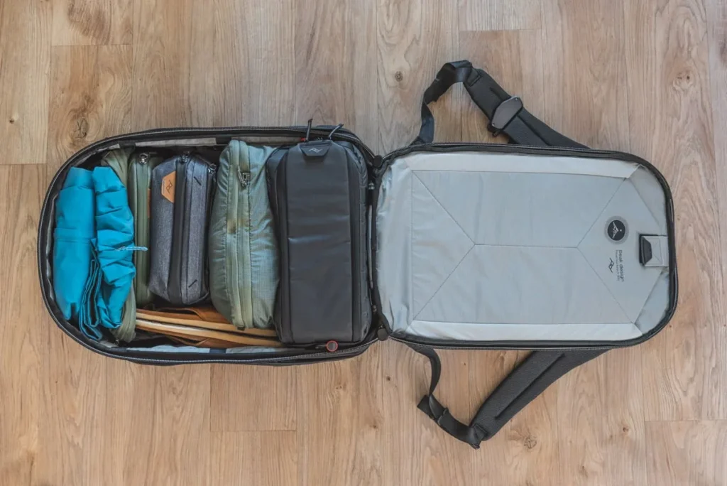 A neatly organized travel kit inside a compact rolling bag. The open suitcase shows multiple small padded pouches and compartments in shades of gray, green, and teal, with folded accessories and a blue packing cube on the left. A light-gray interior panel and black shoulder straps are visible on the right.