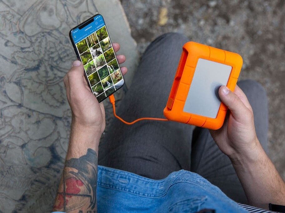 A person sits outdoors, holding a smartphone connected to an orange portable power bank with a built-in SSD-like drive, showcasing power and data backup on the go.