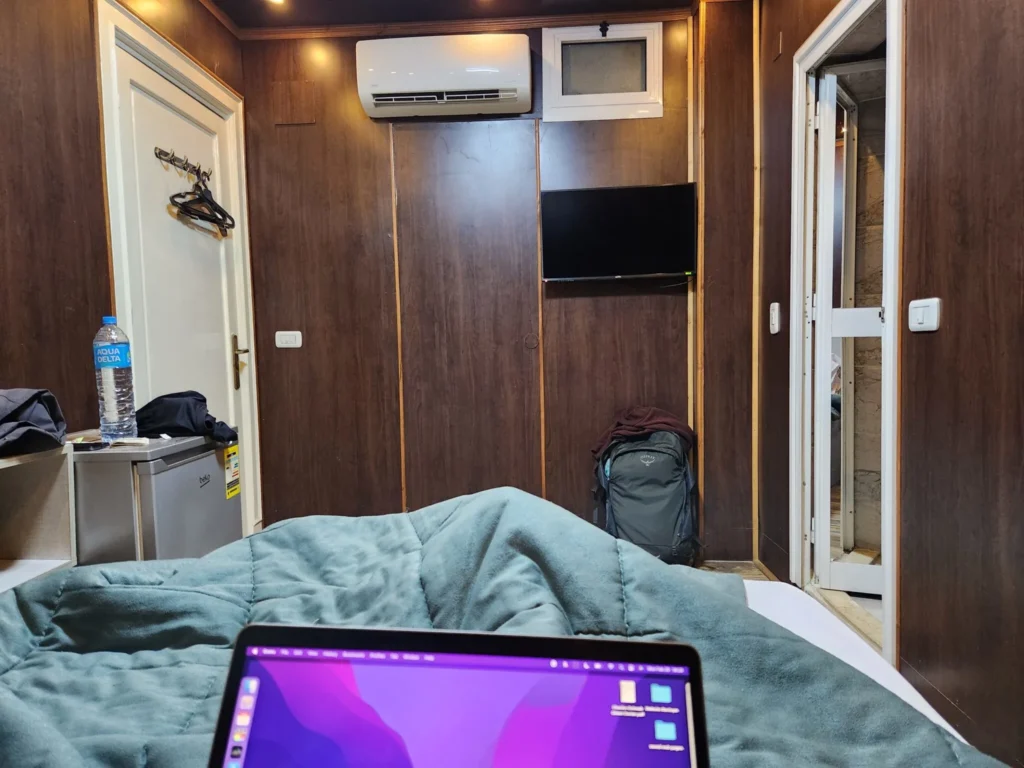 A modern hotel-style hotel room with dark wood paneled walls, a mounted TV, a backpack on the floor, a bottle of water on a mini-fridge, and a laptop resting on a bed with a soft teal comforter in the foreground. An air conditioner is mounted high on the wall, and an open doorway leads to a small bathroom