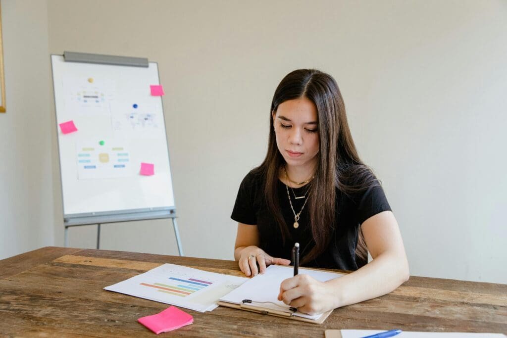 Young woman writing travel video titles in a notebook, with a board of colorful sticky notes behind her.
